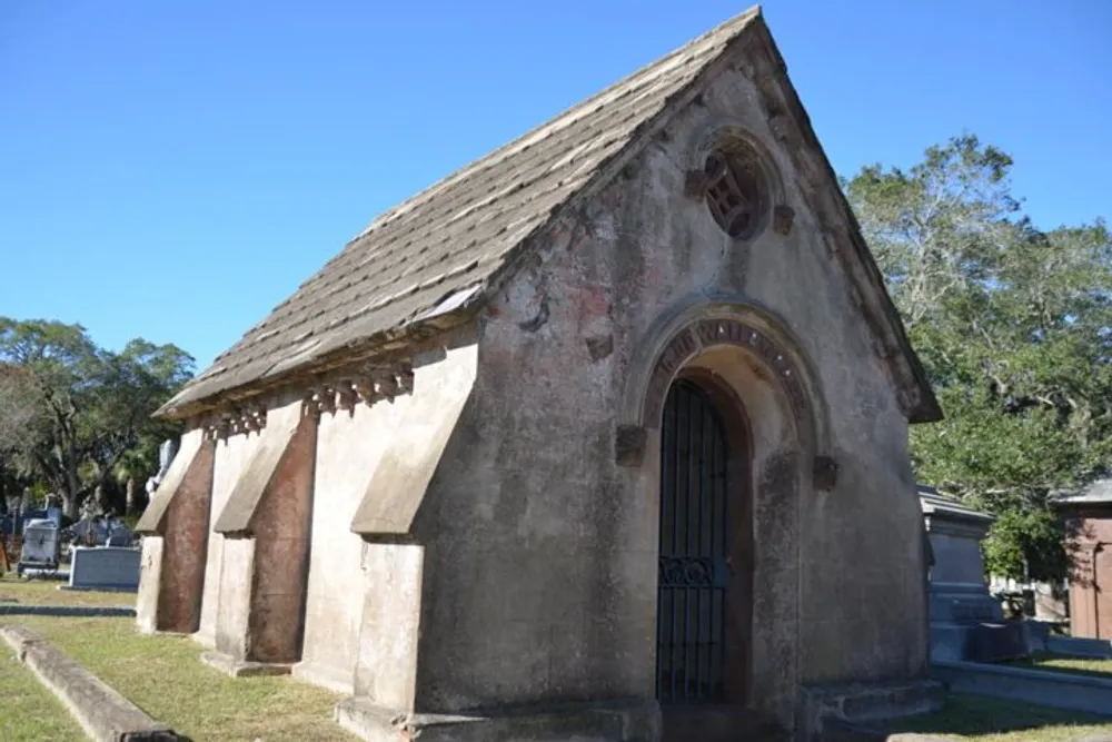 The image shows an old small chapel with a gabled roof in a cemetery under a clear blue sky