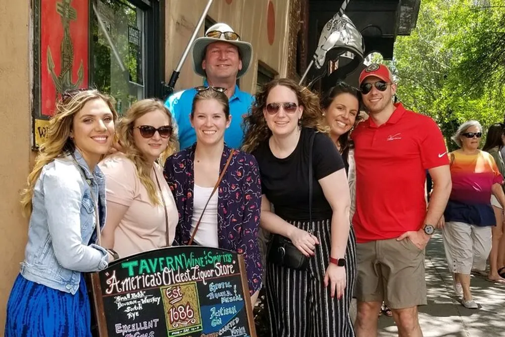 A group of smiling people is posing in front of a sign that reads Tavern Wine  Spirits Americas Oldest Liquor Store Est 1686 with a bustling street scene in the background