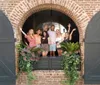 A cheerful group of people are posing and waving with raised hands from a large arched window decorated with plants
