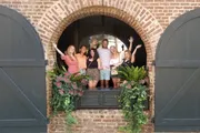 A cheerful group of people are posing and waving with raised hands from a large arched window decorated with plants.