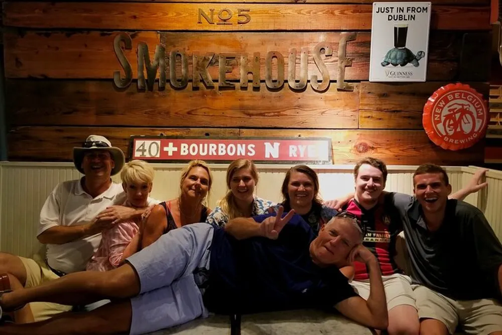 A group of people is posing with smiles in front of a wooden sign that reads Smokehouse with one person playfully lying across the others laps