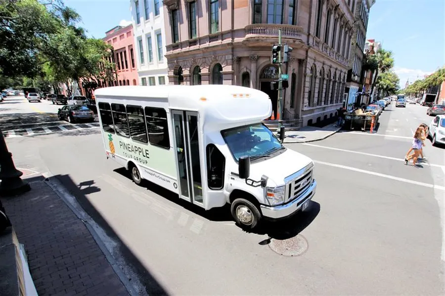 A white shuttle bus with Pineapple Group branding is driving through an urban street with pedestrians and other vehicles nearby.