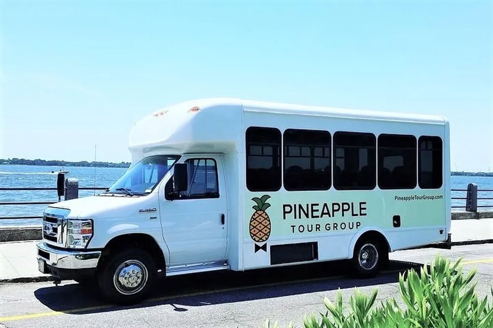 A white shuttle bus with PINEAPPLE TOUR GROUP branding is parked by a waterfront on a sunny day
