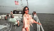 A smiling woman in sunglasses poses on the deck of a boat with other passengers and the American flag fluttering in the background.