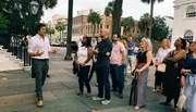 A group of people attentively listens to a man who appears to be giving a tour or a talk outside on a city sidewalk.