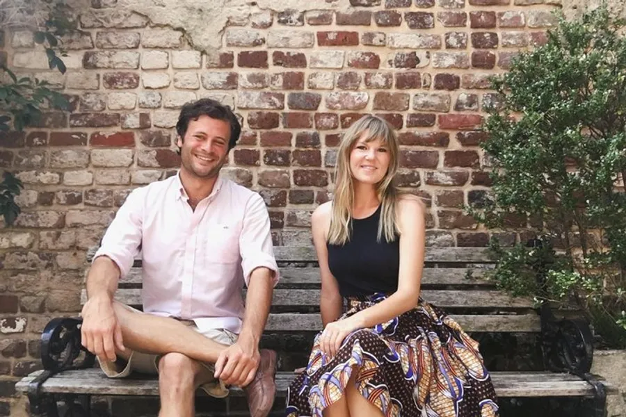 A man and a woman are sitting on a wooden bench, smiling, against a brick wall backdrop with greenery on the right.