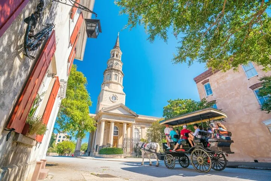 A horse-drawn carriage loaded with passengers tours a historic street with a stately church spire in the background under a clear blue sky.