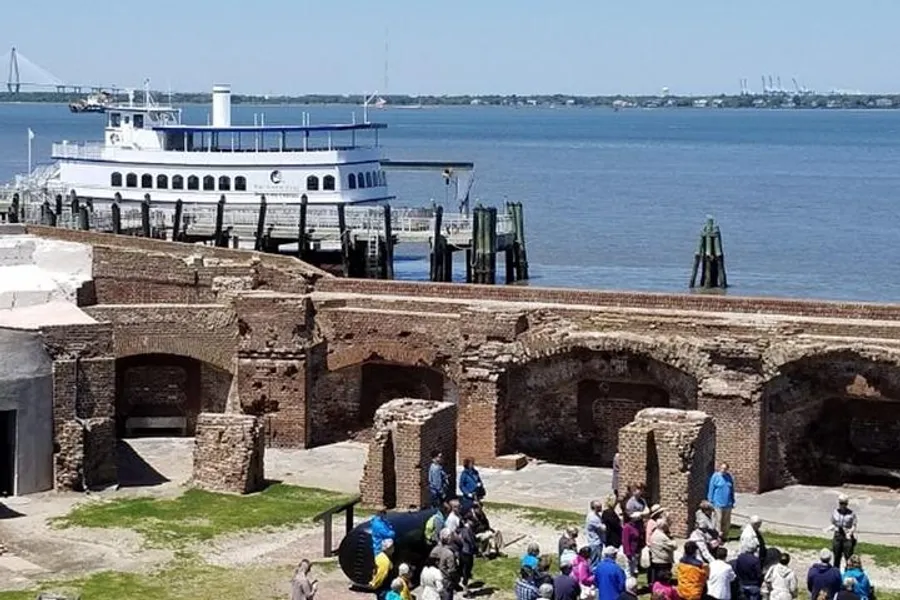This image shows a group of tourists gathered around a cannon display within the ruined walls of an old fort, with a white and blue ferry docked nearby on a bright sunny day.