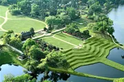 The image shows a lush green landscape with a well-manicured garden, pathways, a body of water, and what appears to be a large estate or historic mansion amidst the foliage.