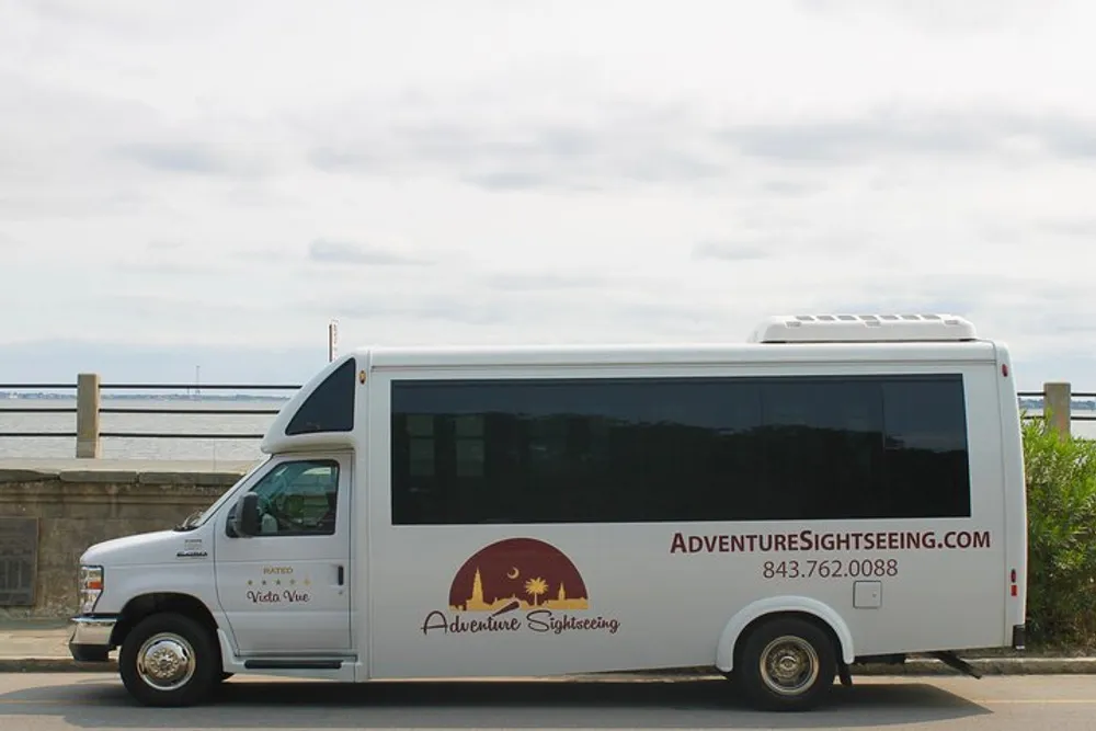 A white sightseeing shuttle bus with the words Adventure Sightseeing and contact information printed on its side is parked near a body of water under a cloudy sky