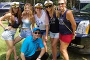 A group of smiling women wearing captain's hats and tops with bridal party themes stands with a man crouching in front of them, all posing for a photo outdoors near a parked vehicle.