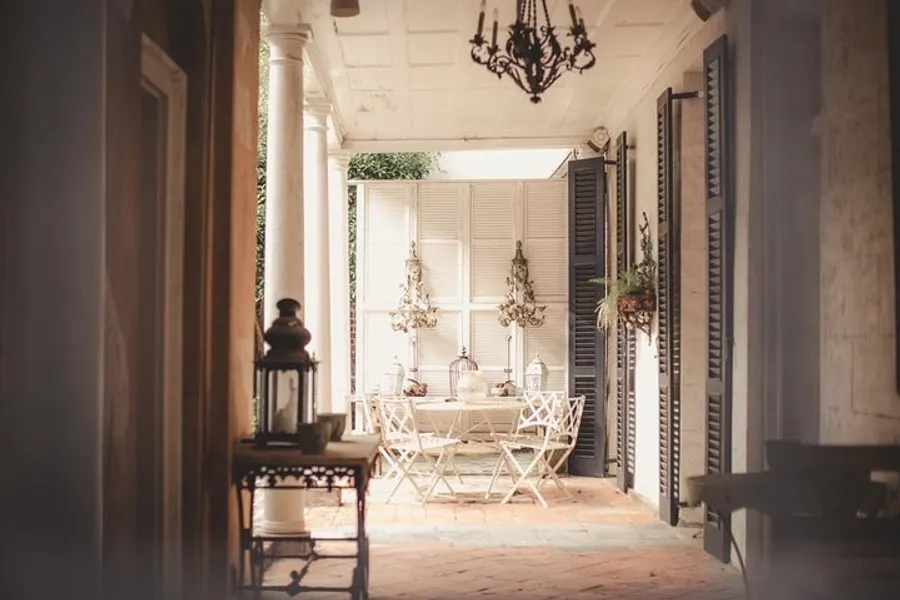 The image shows a quaint, sunlit porch with a vintage aesthetic, featuring wrought iron furniture, hanging plants, and a classic chandelier.