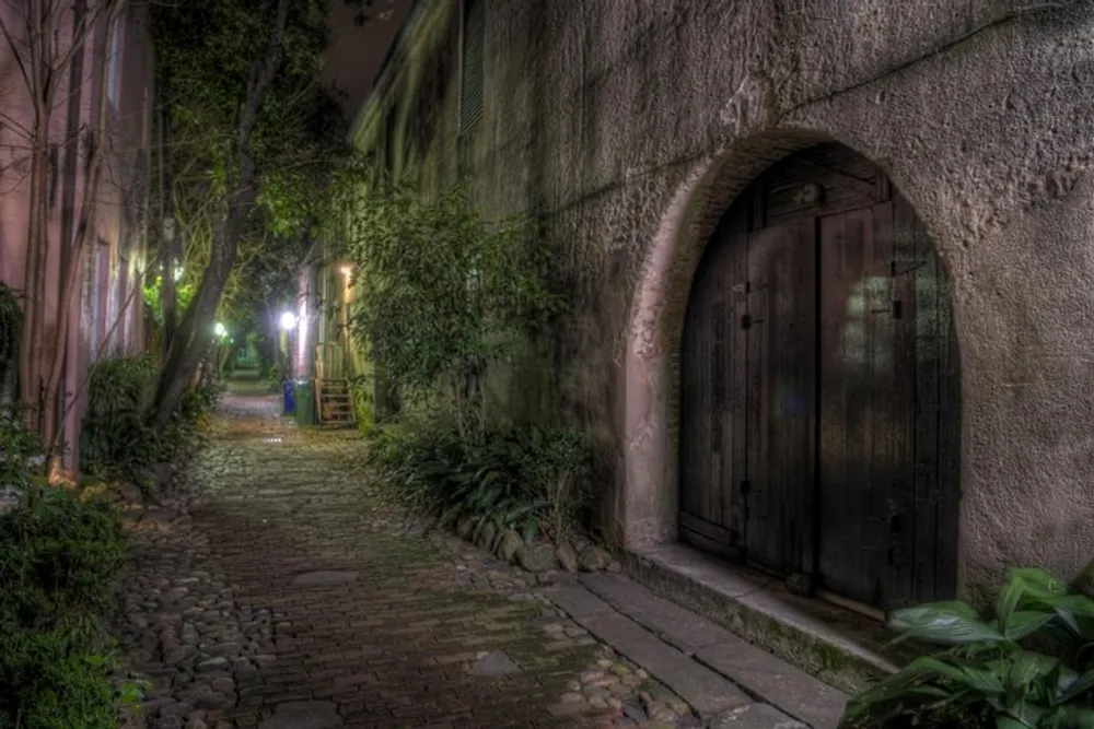 In one sentence The image depicts a mystical narrow cobblestone alley lined with greenery and lit by street lamps at night evoking a sense of old-world charm and mystery