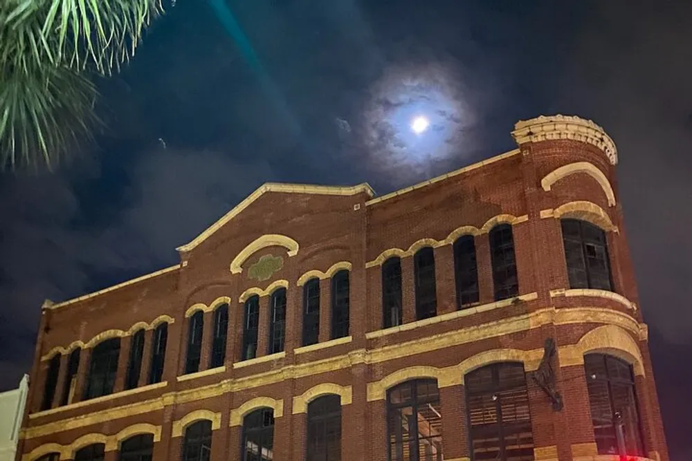 The image shows a historic brick building illuminated at nighttime under a moonlit sky with a palm tree silhouette in the foreground