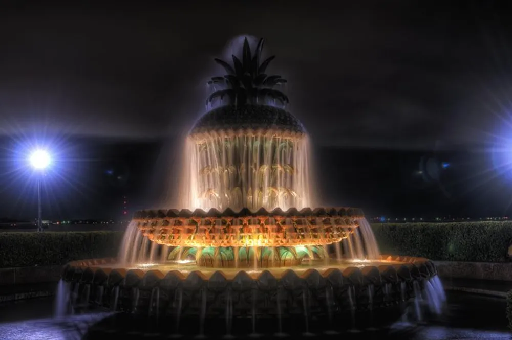 The image depicts a fountain with a pineapple-shaped structure at the top illuminated at night to create a vibrant and dramatic water display