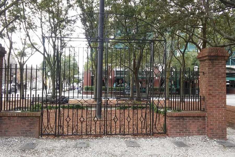 The image shows a black wrought iron fence with ornamental details, mounted on a low brick wall, surrounding a park with trees, benches, and a sidewalk, viewed from a gravel-covered area.