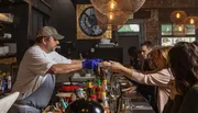 A chef in a casual restaurant is serving a dish to a customer across the bar in a cozy, dimly lit dining atmosphere.