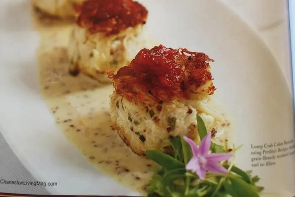 The image shows a magazine page featuring a close-up of two elegantly plated crab cakes topped with a red sauce garnished with herbs and flowers with text indicating its from CharlestonLivingMagcom