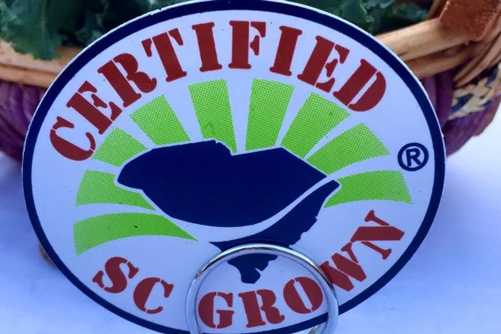 The image shows a close-up of a keychain featuring a logo with the text CERTIFIED SC GROWN around a graphic depicting two hands cradling a sunburst pattern