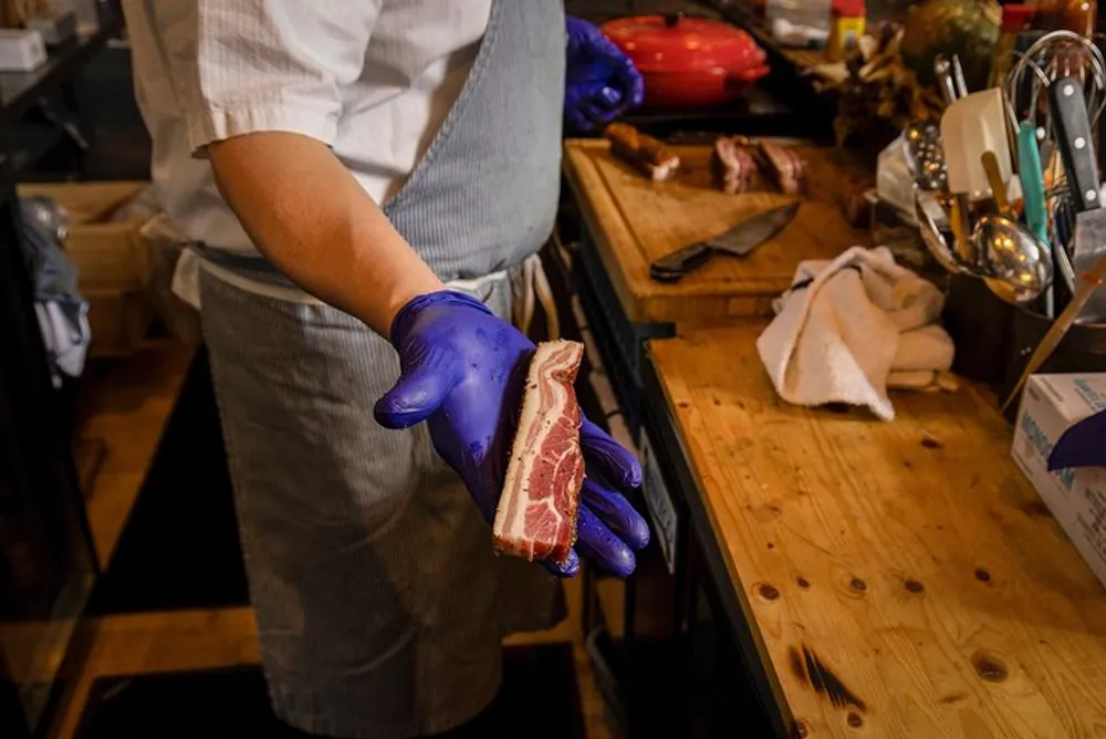 A person wearing a blue glove and an apron is holding a raw cut of meat in a kitchen environment