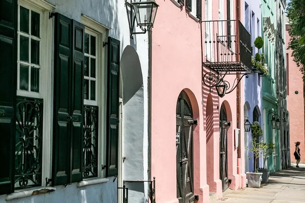 The image shows a charming row of pastel-colored buildings with traditional shutters wrought-iron details and a pedestrian strolling along the sunlit sidewalk