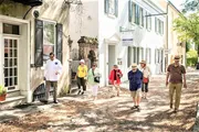 A group of tourists, led by a guide, is walking down a picturesque urban cobblestone street lined with historic buildings on a sunny day.
