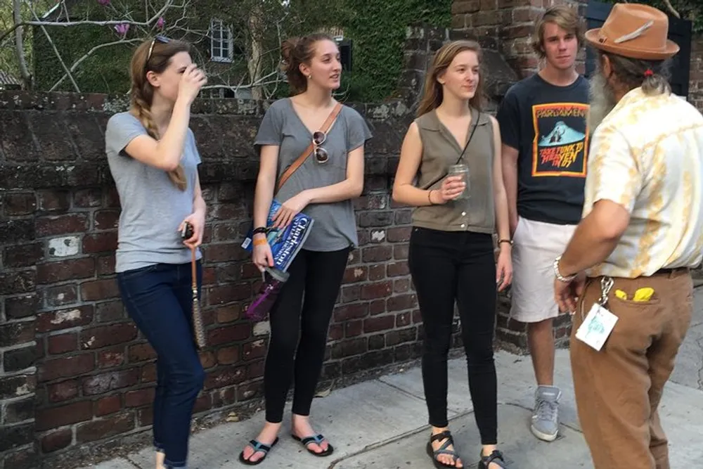 A group of young adults is engaged in a casual conversation with an older individual on a sidewalk next to a brick wall
