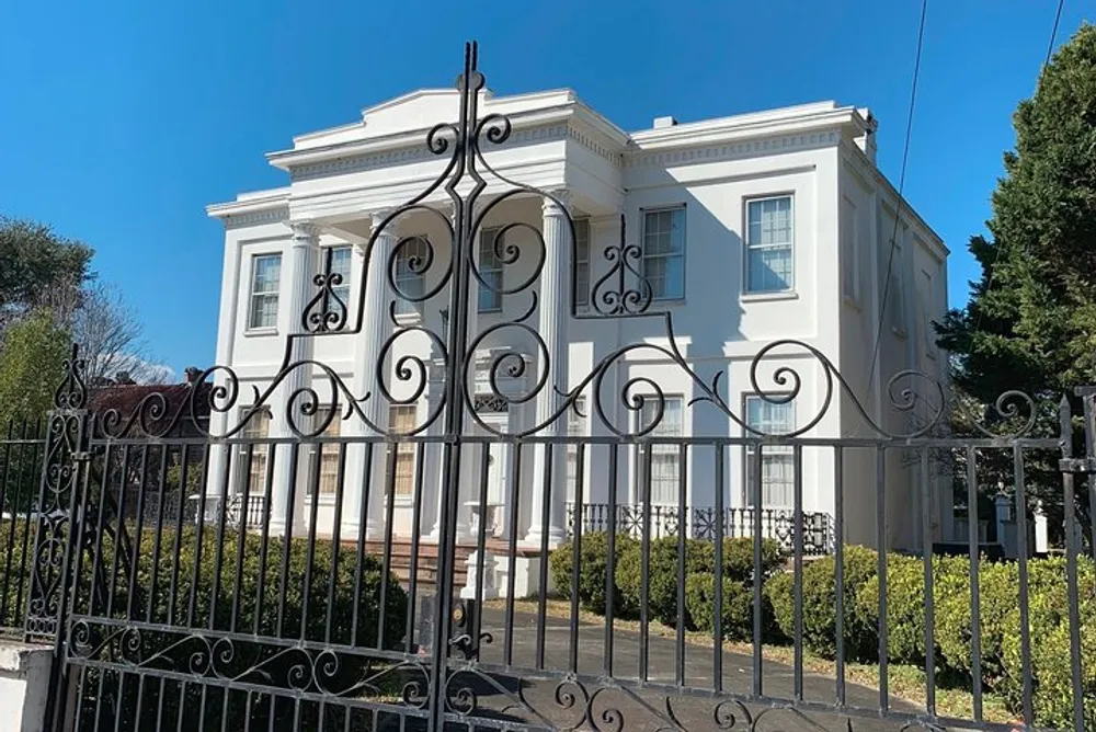 An elegant white house with classic architectural features is enclosed behind ornate black wrought iron gates under a clear blue sky