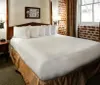 The image shows a neatly made bed in a well-lit hotel room with exposed brick walls and traditional decor