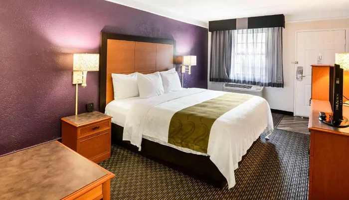 The image shows a neatly arranged hotel room with a double bed decorative headboard and modern amenities like a flat-screen TV and air conditioner
