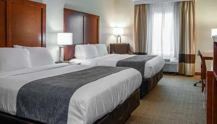 The image shows a well-kept clean hotel room featuring two double beds with white linens and dark runners flanked by nightstands with lamps set against a backdrop of earth-tone curtains by a window and a neutral-toned carpeted floor