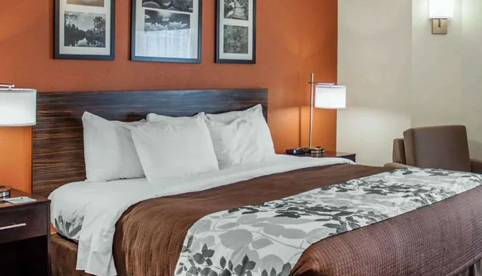 The image shows a neatly made hotel room with a large bed crisp white bedding a floral patterned throw and warm-toned walls flanked by artwork and modern lighting