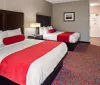 The image shows a tidy hotel room with two beds featuring white linens and red accent pillows and runners along with a nightstand desk and an open bathroom door in the background