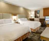 The image shows a neatly arranged hotel room with two double beds a flat-screen TV lamps and simple decor