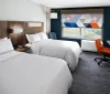 The image shows a modern hotel room with two double beds a work desk with an orange chair and a window with a view of greenery outside