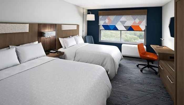 The image shows a modern hotel room with two double beds a work desk with an orange chair and a window with a view of greenery outside