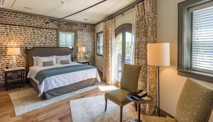 The image depicts a cozy and inviting bedroom featuring a bed with a classic headboard against an exposed brick wall accented by elegant furnishings and natural light streaming in through the windows