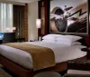 The image shows a modern and neatly arranged hotel room with a large bed a work desk with a laptop a sitting area and a large framed picture of a propeller airplane on the wall
