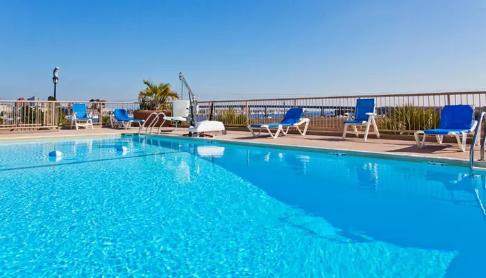 A clear blue swimming pool on a sunny day with deck chairs lined up along the poolside overlooking a beach or ocean horizon