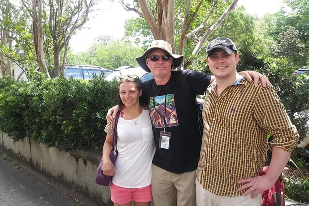 Three people are smiling for a photo outdoors on a sunny day with trees and parked cars in the background