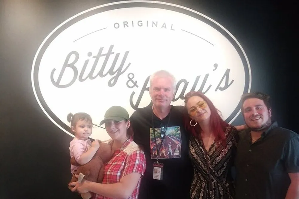 Four people are posing for a photo in front of a wall with a logo that reads Bitty  Beaus including an adult holding a baby an adult male and two other adults one of whom is wearing eyeglasses