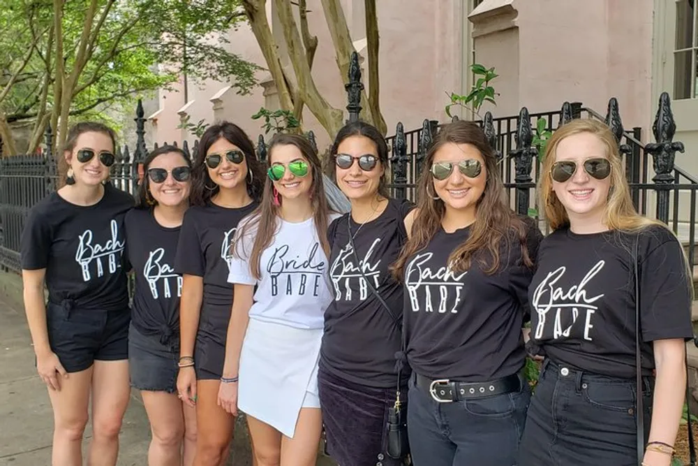 The image features a group of smiling women wearing matching black t-shirts with Bach Babe written on them except for one in the center wearing a white Bride Babe t-shirt likely celebrating a bachelorette party