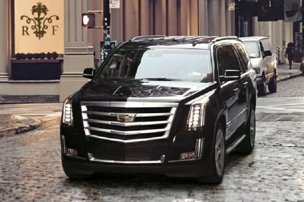 A black Cadillac SUV is driving on a cobblestone street with other cars in the background illustrating an urban setting
