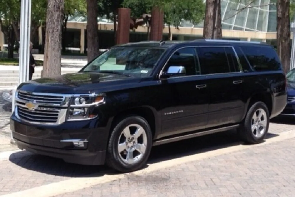 The image shows a black Chevrolet Suburban parked on a sunny street beside a sidewalk lined with trees