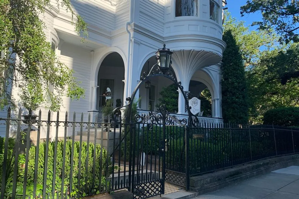The image shows the ornate entrance of a grand white house with a curved bay window wrought iron gate and lush greenery