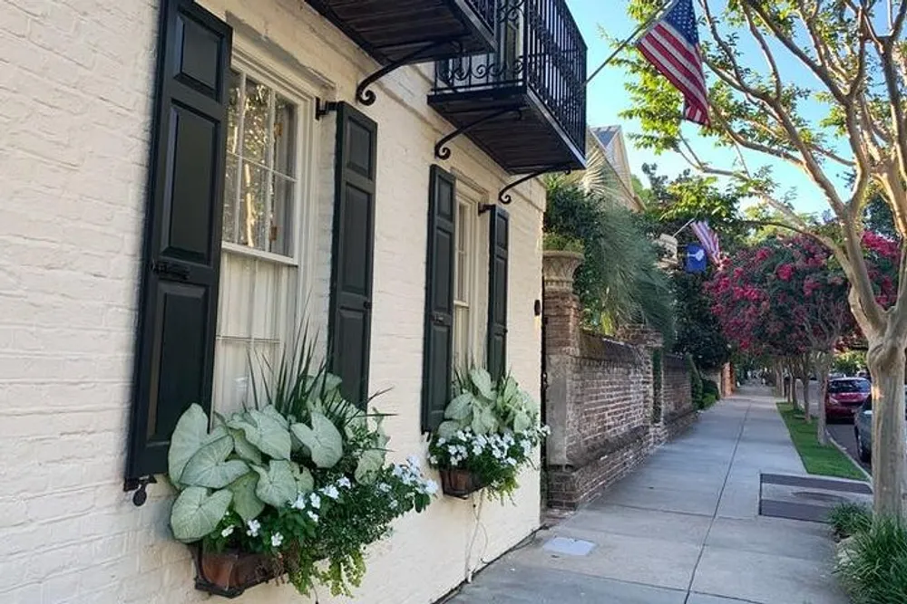 This image shows a charming street scene with a cream-colored building adorned with green window shutters flower boxes and an American flag flanked by a brick sidewalk and lush greenery
