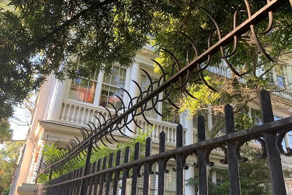 An ornate iron fence with curved spikes tops the barrier in front of a classic white house with large windows and columns partially obscured by lush green foliage