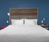 The image shows a modern bedroom with a large bed white bedding a dark blue wall a wooden headboard and minimalist decor including a standing lamp and bedside table with a lamp