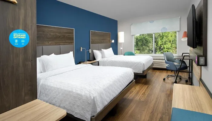 The image shows a modern hotel room with two beds a work area a mounted TV and a large window letting in natural light