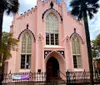 A pastel pink Gothic Revival style church features pointed arch windows and a welcoming banner by the entrance framed by a palm tree and wrought iron fence under a blue sky with scattered clouds